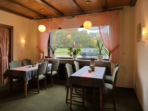 Innenansicht Restaurant - Haus Koppelberg Gemütliches Gasthauszimmer mit Holzbalkendecke, zwei gedeckten Tischen und großem Fenster mit Naturblick.