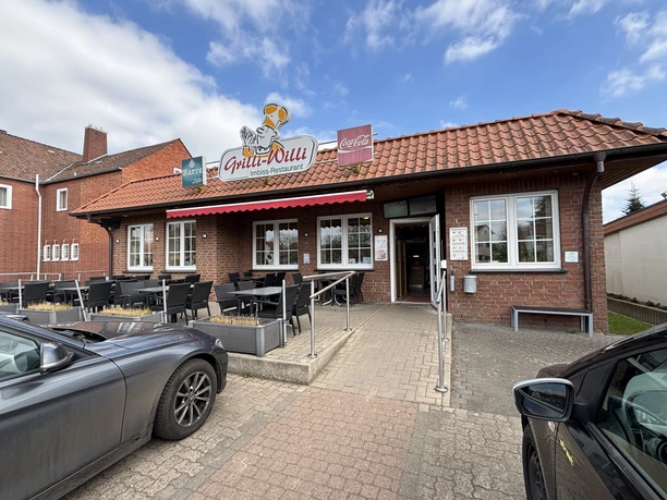 Steinernes Gebäude mit Terrasse, roten Dachziegeln und Gastronomieschild, neben parkenden Autos.