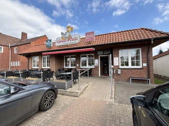 Grilli-Willi Steinernes Gebäude mit Terrasse, roten Dachziegeln und Gastronomieschild, neben parkenden Autos.