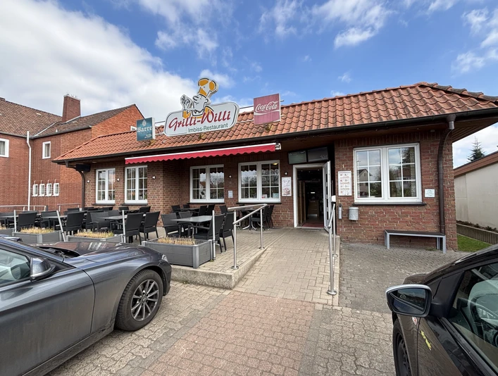 Grilli-Willi Steinernes Gebäude mit Terrasse, roten Dachziegeln und Gastronomieschild, neben parkenden Autos.