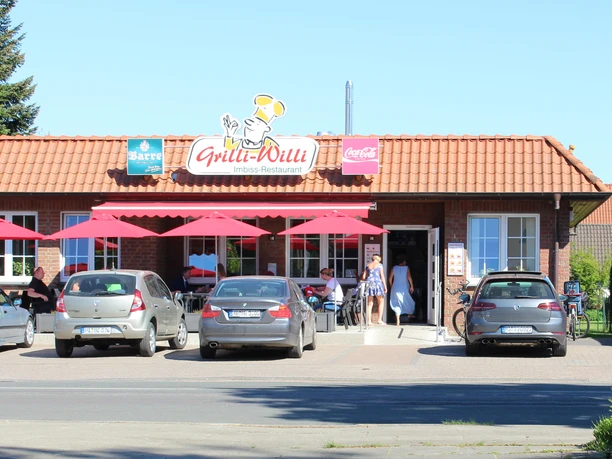 Grilli-Willi Gemütliches Imbiss-Restaurant mit rotem Dach, roten Sonnenschirmen und parkenden Autos davor.