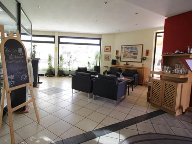 Heller Eingangsbereich mit Sitzgruppen, Theke, Pflanzen und Informationstafel nahe der Glastür