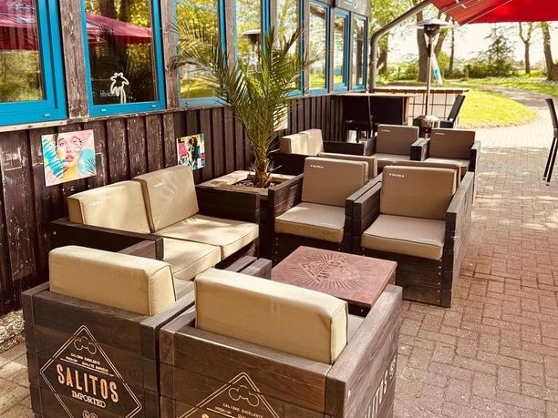 Beachbar 12 am Badesee Lahde Außenbereich einer Strandbar mit Holzmöbeln, Schildern und Sonnenschirmen nahe eines Badesees.