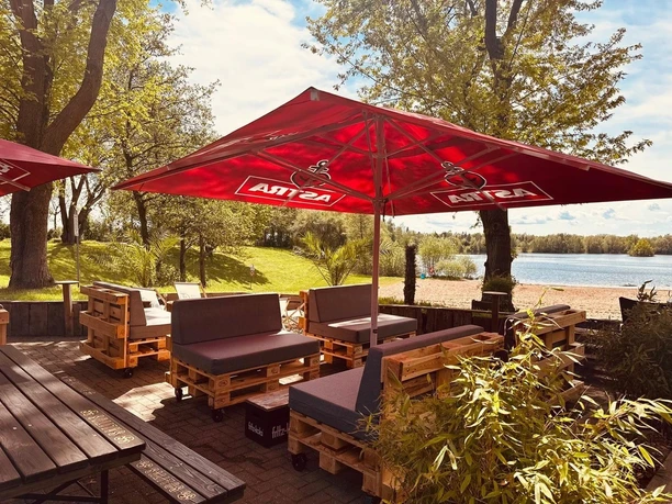 Beachbar12 am Badesee Lahde Sitzgruppe aus Palettenmöbeln unter roten Sonnenschirmen, umgeben von Bäumen nahe einem Badesee.