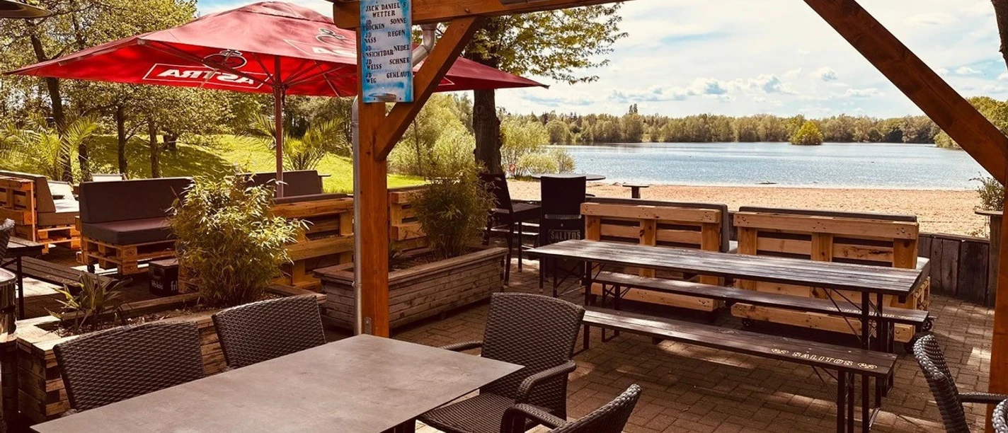Beachbar12 am Badesee Lahde Terrasse einer Beachbar mit Holzbelag, Blick auf einen Badesee, sonniger Himmel, rote Sonnenschirme.