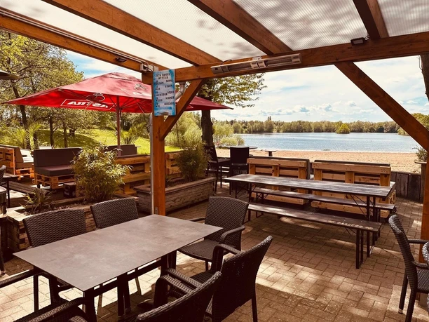 Beachbar12 am Badesee Lahde Terrasse einer Beachbar mit Holzbelag, Blick auf einen Badesee, sonniger Himmel, rote Sonnenschirme.