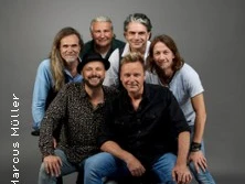 Sechs Musiker posieren lächelnd vor grauem Hintergrund. Vier sitzen, zwei stehen dahinter.