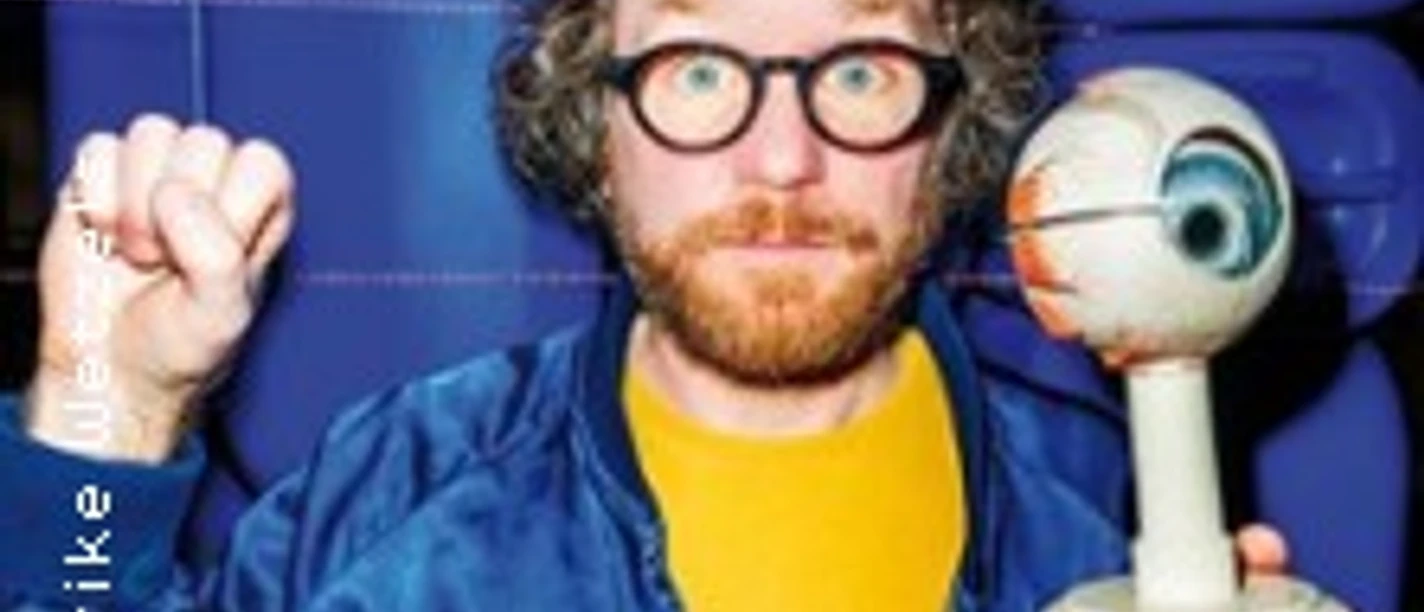 Ein Mann mit Brille hält ein Augenmodell in der Hand, im Hintergrund blaue Sitzreihen.