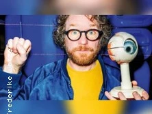 Ein Mann mit Brille hält ein Augenmodell in der Hand, im Hintergrund blaue Sitzreihen.