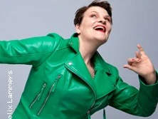 Frau in grüner Lederjacke mit ausdrucksvollem Gesichtsausdruck vor einfachem, grauem Hintergrund.