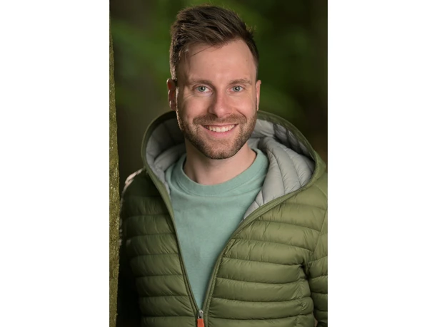 wanderung.jpg Lächelnder Mann in grüner Jacke lehnt im Wald an einem Baumstamm.