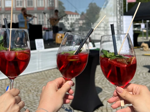 Weinfest_Weingläser.jpeg 3 Menschen halten Weingläser mit roten Getränken. Im Hintergrund spielen Musiker.