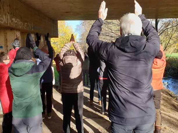 Personengruppe bei Gymnastikübungen im Freien unter Brücke, bunt gekleidet, herbstliche Atmosphäre.