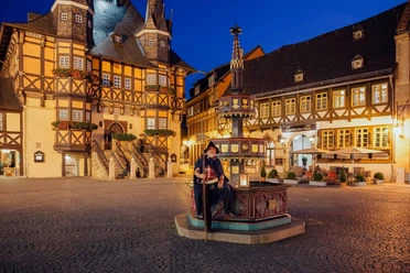 Zu sehen ist ein als Nachtwächter verkleideter Mann, der auf dem Brunnen vor dem nächtlichen Rathaus Wernigerode sitzt.