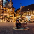 gothisches-haus-wernigerode-rahmenprogramm-nachtwaechterrundgang.jpg Zu sehen ist ein als Nachtwächter verkleideter Mann, der auf dem Brunnen vor dem nächtlichen Rathaus Wernigerode sitzt.