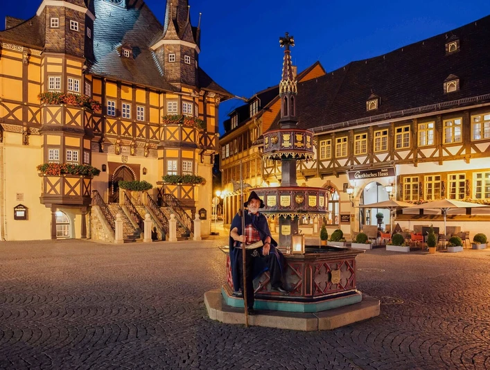 gothisches-haus-wernigerode-rahmenprogramm-nachtwaechterrundgang.jpg Zu sehen ist ein als Nachtwächter verkleideter Mann, der auf dem Brunnen vor dem nächtlichen Rathaus Wernigerode sitzt.