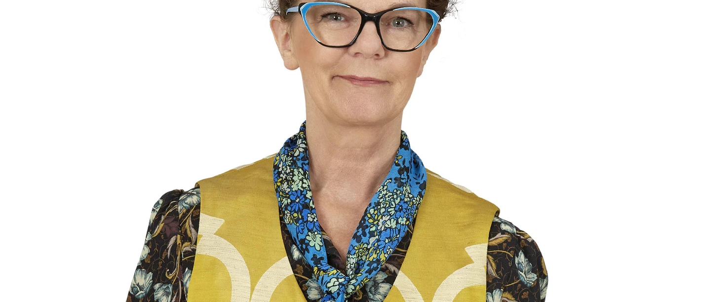 Eine Frau mit modischer Brille, gemustertem Kleid und Halstuch vor weißem Hintergrund.