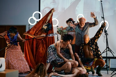 An ensemble of actors is grouped in theatrical, gesturing poses around a person lying on the stage floor.