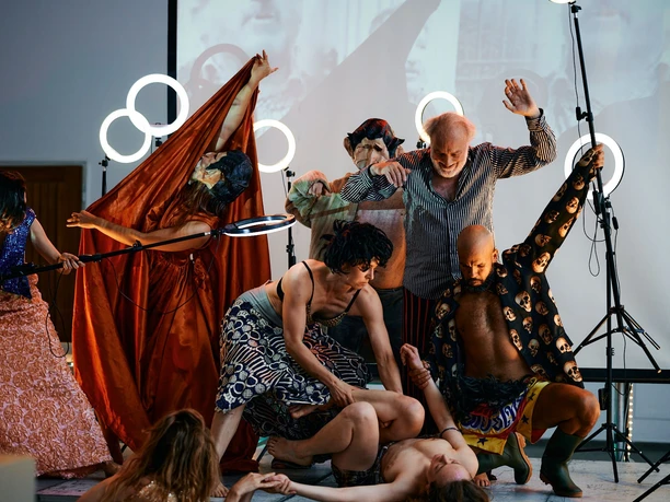 An ensemble of actors is grouped in theatrical, gesturing poses around a person lying on the stage floor.