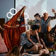 SommerBlut4(c)Nathan Dreessen.jpg Ein Ensemble von Schauspieler*innen ist in theatralen, gestikulierenden Posen um eine am Boden liegende Person auf einer Theaterbühne gruppiert.