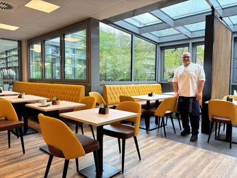 Cafe-am-Rosengarten-Betreiber-Can-Bentami.jpg Heller Innenraum vom Café am Rosengarten mit großen Fenstern. Der freundliche Betreiber steht neben eleganten Möbeln in warmen Tönen.