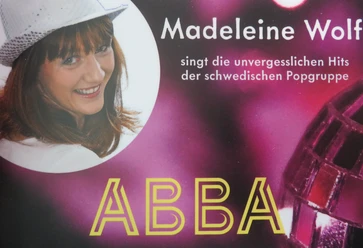 Madeleine Wolf smiles in a glittering hat as she sings ABBA hits on a colorful stage.