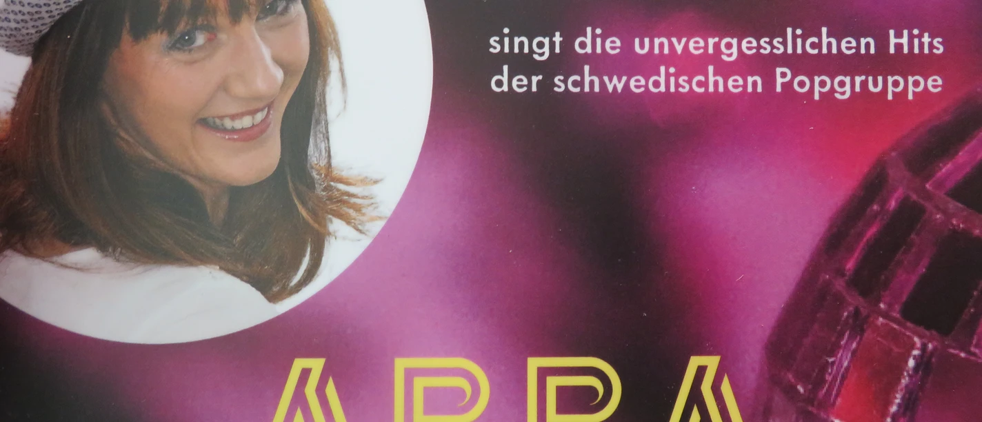 Madeleine Wolf - ABBA Madeleine Wolf lächelt in glitzerndem Hut, während sie ABBA-Hits auf einer bunten Bühne singt.