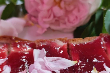Close-up of a loaf of bread with red jam and pink petals on it, a blurred pink flower in the background.