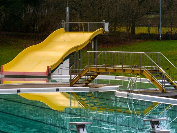 Gelbe Rutsche im Freibad mit stufenlosem Zugang und spiegelndem Pool, umgeben von Bäumen und Grünflächen.