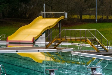 20010-FreibadRutsche_©GesUndTourismus Horn-Bad Meinberg GmbH.jpg Gelbe Rutsche im Freibad mit stufenlosem Zugang und spiegelndem Pool, umgeben von Bäumen und Grünflächen.