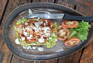 Essen Ein rustikaler Holztisch mit einem Teller Garnelen-Avocado-Toast, garniert mit Feta und Salat.