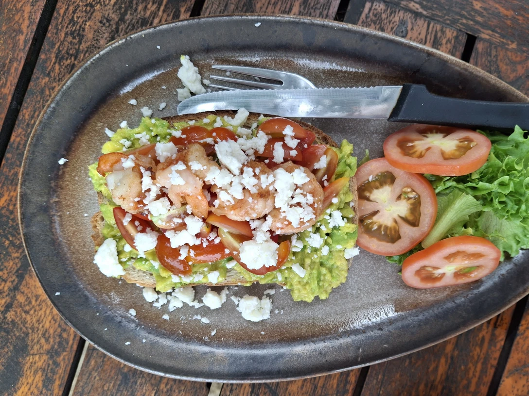 Essen Ein rustikaler Holztisch mit einem Teller Garnelen-Avocado-Toast, garniert mit Feta und Salat.A rustic wooden table with a plate of prawn and avocado toast, garnished with feta and salad.Et rustikt træbord med en tallerken reje- og avocadotoast, pyntet med feta og salat.Een rustieke houten tafel met een bord garnalen- en avocadotoast, gegarneerd met feta en salade.