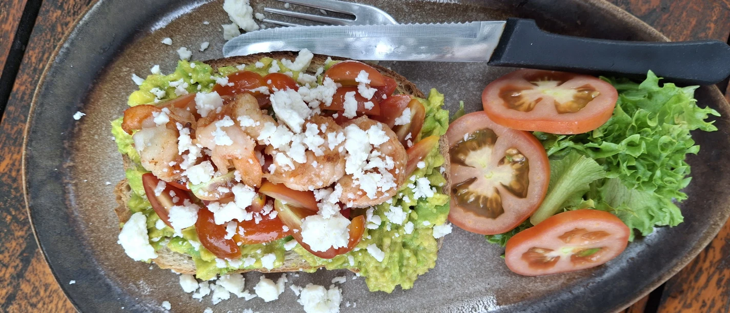 Essen Ein rustikaler Holztisch mit einem Teller Garnelen-Avocado-Toast, garniert mit Feta und Salat.
