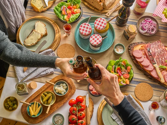 Gedeckter Tisch mit TYPISCH-neanderland Produkten und Ratuga Bier Einladender Tisch mit regionalen Produkten, Salaten und Ratuga Bier, Hände beim Anstoßen.