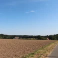 Feldweg führt an weitläufigen Ackerflächen vorbei, im Hintergrund sind bewaldete Hügel zu sehen.