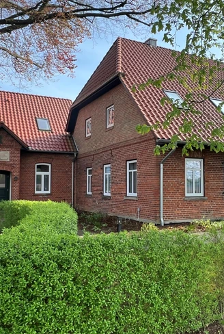 Backsteingebäude mit roten Dachziegeln und vielen Fenstern, umgeben von gepflegtem Grün und Bäumen.Brick building with red roof tiles and many windows, surrounded by well-tended greenery and trees.Murstensbygning med røde tagsten og mange vinduer, omgivet af velplejede grønne områder og træer.Bakstenen gebouw met rode dakpannen en veel ramen, omgeven door goed onderhouden groen en bomen.