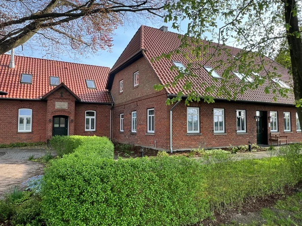 Bakstenen gebouw met rode dakpannen en veel ramen, omgeven door goed onderhouden groen en bomen.