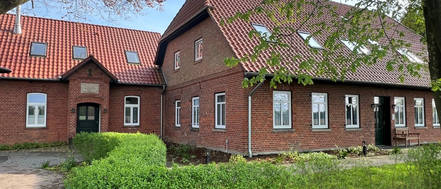 Alte Schule Kirchlinteln Brick building with red roof tiles and many windows, surrounded by well-tended greenery and trees.