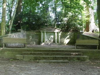 POI_2-Familiengrab.JPG Zwei Holzsitzbänke flankieren ein steinernes Familiengrab in einem von Bäumen gesäumten Waldstück.