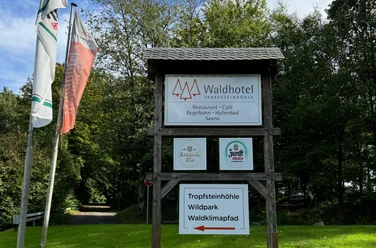 Schild - Waldhotel Tropfsteinhöhle Waldhotel-Schild mit Angeboten wie Restaurant, Kegelbahn und Sauna vor grünen Bäumen und Flaggen.