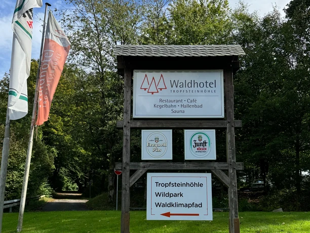 Schild - Waldhotel Tropfsteinhöhle Waldhotel-Schild mit Angeboten wie Restaurant, Kegelbahn und Sauna vor grünen Bäumen und Flaggen.