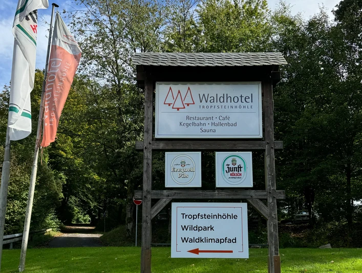 Schild - Waldhotel Tropfsteinhöhle Waldhotel-Schild mit Angeboten wie Restaurant, Kegelbahn und Sauna vor grünen Bäumen und Flaggen.