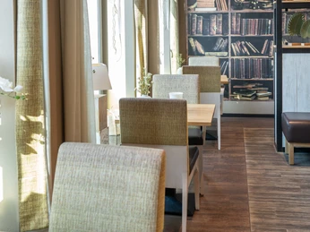 Ferdinands Bäckerei | Restaurant Ein sonnendurchfluteter Innenraum mit Sitzgarnituren und einem dekorativen Bücherregal an der Wand.