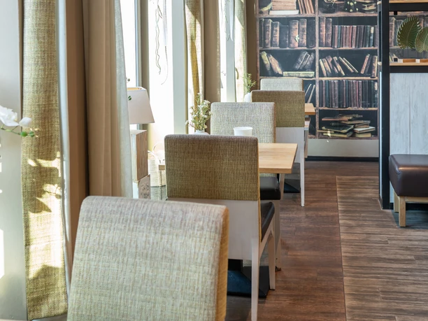 Ferdinands Bäckerei | Restaurant Ein sonnendurchfluteter Innenraum mit Sitzgarnituren und einem dekorativen Bücherregal an der Wand.