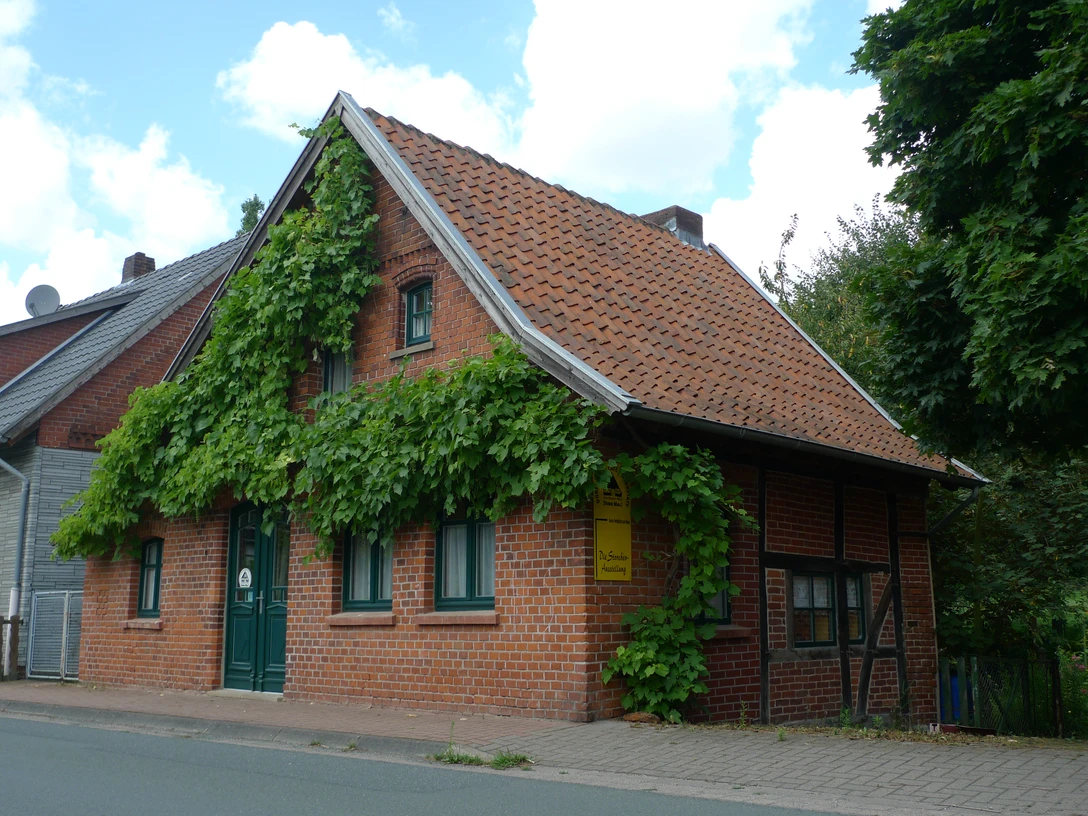 Dat lüttke Hues Backsteinhaus mit Satteldach und grüner Fassadenbewachsung an einer Straßenkreuzung, bewölkter Himmel.
