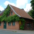 Dat lüttke Hues Backsteinhaus mit Satteldach und grüner Fassadenbewachsung an einer Straßenkreuzung, bewölkter Himmel.