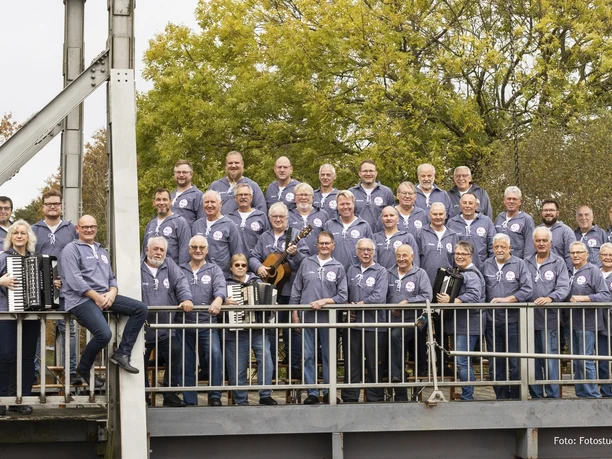 Fehntjer Shanty-Chor Eine große Gruppe älterer Männer in maritimen Outfits, einige mit Akkordeons, steht lächelnd auf einer Brücke. Der goldene Herbsthintergrund ergänzt den Freiluftauftritt des Fehntjer Shanty-Chors.