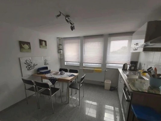 Ferienwohnung Angelmann Moderne Küche mit Esstisch, sechs Stühlen, Fensterjalousien und diversen Küchengeräten.