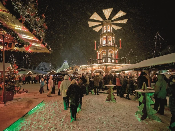 Ein festlicher Weihnachtsmarkt bei Nacht, beleuchtet von bunten Lichtern. Menschen genießen die Atmosphäre im Schnee um eine große Drehscheibe.