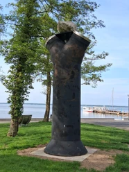 Sculpture made of dark metal on green grass in front of trees and lake. There is a jetty in the background.